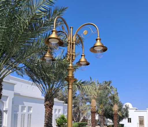 Rijk versierde straatlantaarn in gouden kap met vier 04/Aries/A armaturen, tegen een achtergrond van palmbomen en witte gebouwen op een zonnige dag